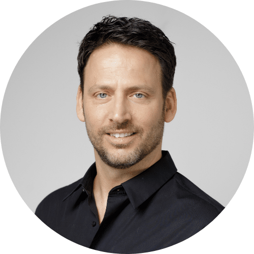 Man with short dark hair and beard wearing a black button-up shirt, posing and smiling slightly against a plain light background.