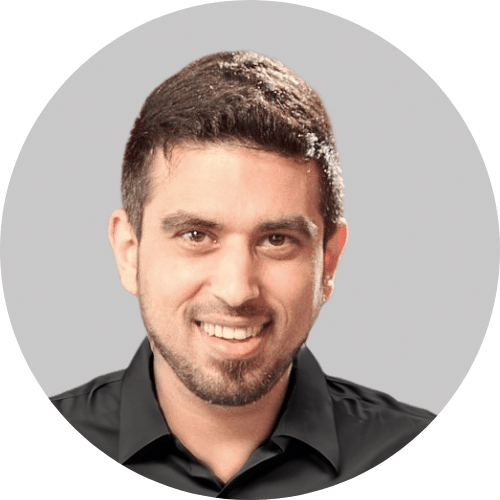 A man with short dark hair and a beard, wearing a black collared shirt, smiles at the camera against a plain gray background.