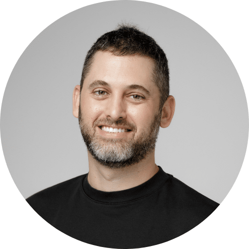 A man with short dark hair and a beard, wearing a black shirt, smiling at the camera against a plain light background.