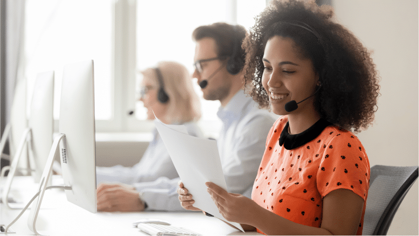 Three customer service representatives wearing headsets sit at desks with computers. The woman in the foreground is holding papers and smiling.