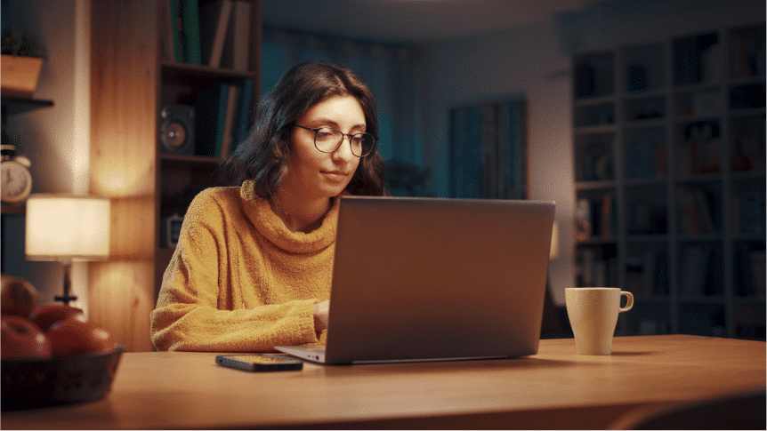 A person with glasses sits at a wooden table, using a laptop with a mug and smartphone nearby, in a warmly lit room with shelves and a lamp in the background.