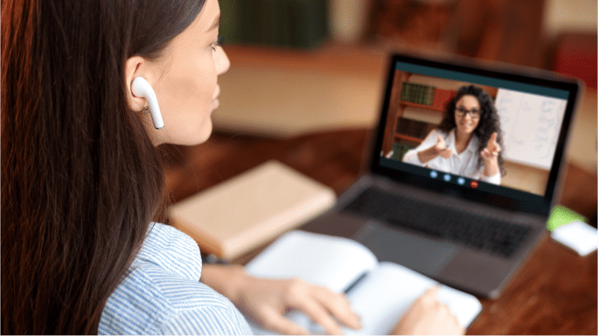 A woman wearing wireless earbuds sits at a desk with an open notebook, attending a video call with another woman displayed on her laptop screen.