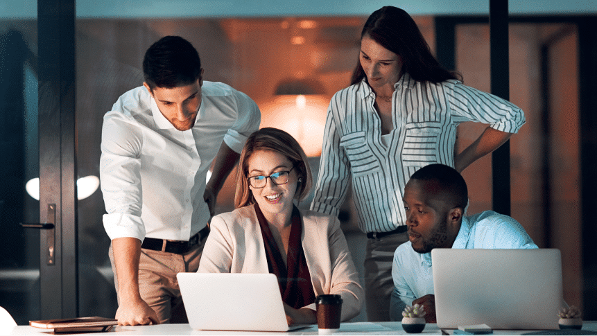 Four people in business attire gather around a desk, focused on a laptop screen, working together in a modern office setting.