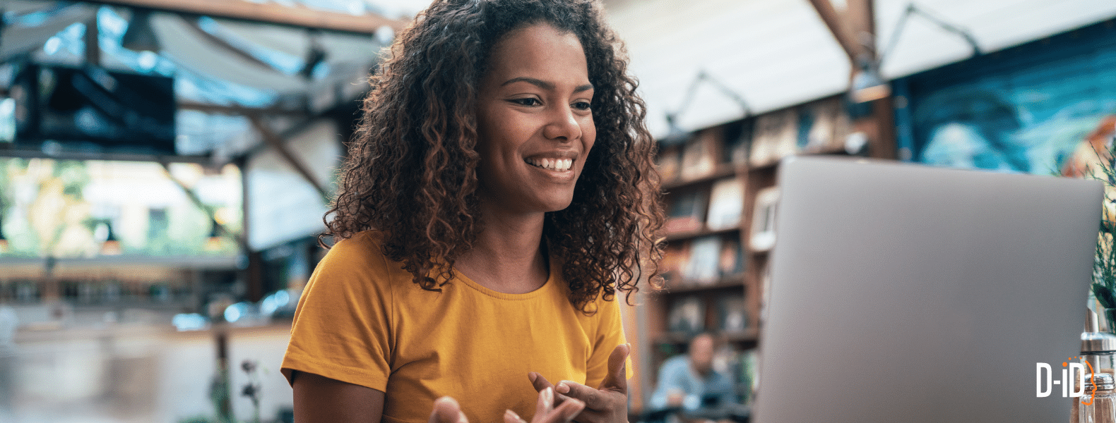 A woman with curly hair in a yellow shirt smiles while video chatting on a laptop in a bright, modern indoor space—showcasing personalized video content. The DID logo appears in the bottom right corner.