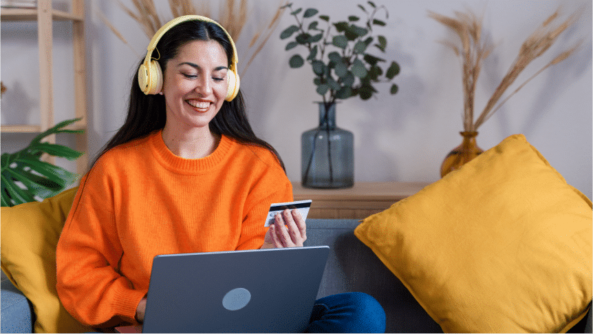 Woman wearing yellow headphones and orange sweater sits on a sofa, holding a credit card and using a laptop, with yellow cushions and decorative plants in the background.