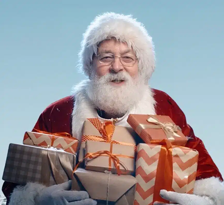 A man dressed as Santa Claus holds a stack of wrapped presents with various ribbons, standing against a plain blue background.