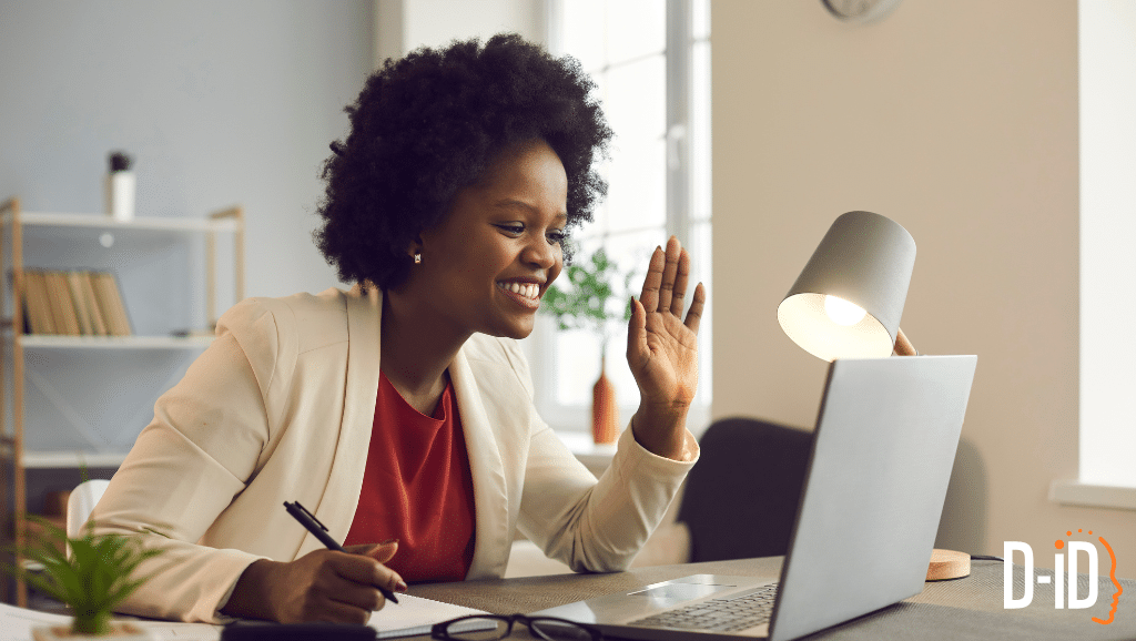 A woman sits at a desk with a notebook, smiling and waving at her laptop during a video call powered by a conversational AI platform in a cozy home office setting.